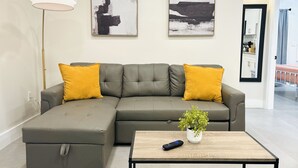 A modern living room with a sectional sofa, two mustard-colored pillows, a small table with a remote control, and a potted plant. - Cute & Spacious 2BR/2Bath Home in FLL w/ Patio (Fort Lauderdale)