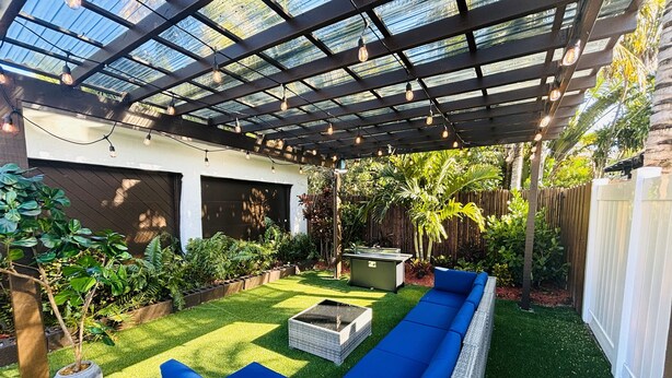 A covered patio area with a long blue sofa, a fire pit, and various plants. - Cute & Spacious 2BR/2Bath Home in FLL w/ Patio (Fort Lauderdale)