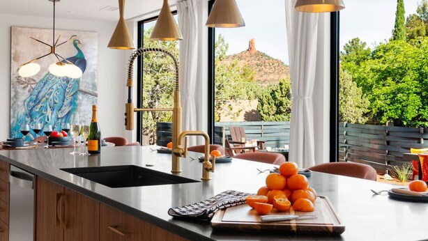 A modern kitchen with a large island, pendant lights, a peacock painting, and a view of greenery and hills.