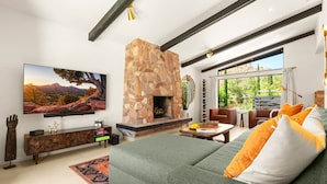 A modern living room with a stone fireplace, a large flat-screen TV, a comfortable sofa, and a view of greenery through large windows.