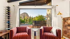 A modern living room with two red velvet armchairs, a small round table with a glass of wine, a bookshelf, and a large window with a mountain view.
