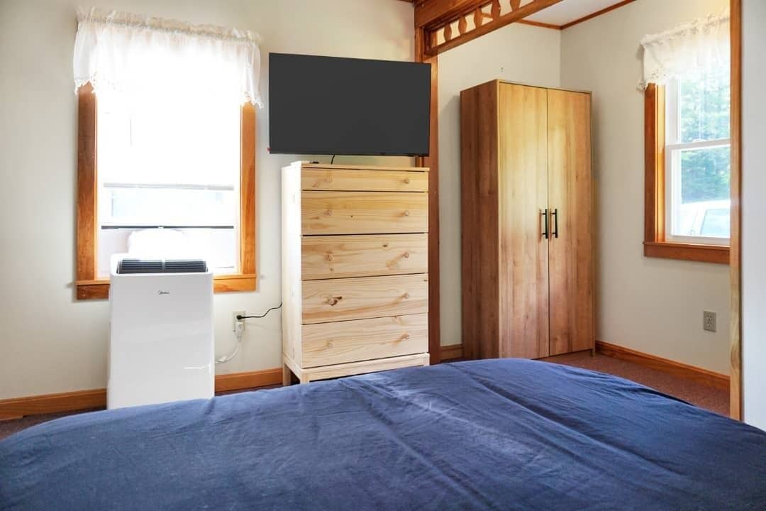 A bedroom with a bed, a wooden wardrobe, a TV mounted on the wall, and a window with curtains.