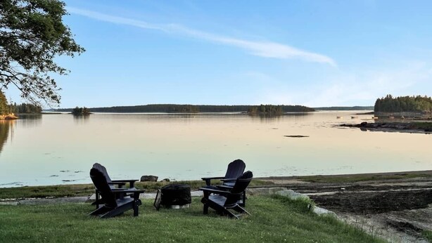 A lakeside view with two Adirondack chairs and a small table, surrounded by grassy areas and trees. - Oceanfront Cottage • Firepit + Beach • Near Acadia & Bar Harbor  (Milbridge)
