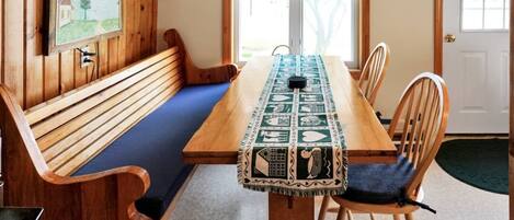 Una mesa de comedor de madera con mantel estampado, rodeada de sillas de madera con asientos acolchados. Una banca se encuentra a lo largo de la pared, y un cuadro enmarcado cuelga sobre la mesa.