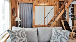A cozy living room with a grey sofa, patterned cushions, a wooden staircase, and a window with blinds. - Oceanfront Cottage • Firepit + Beach • Near Acadia & Bar Harbor  (Milbridge)