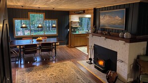 A cozy interior with a wooden dining table, chairs, a fireplace, and a large window overlooking a forest. - Naked Elk Lodge: 4 Bdrms, Hot Tub, Projector TV near Mt. Rainier (Packwood)