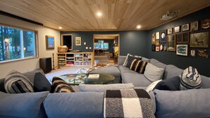 A modern living room with a large sectional sofa, a coffee table, and a bookshelf. There is a large window providing a view of the outdoors. - Naked Elk Lodge: 4 Bdrms, Hot Tub, Projector TV near Mt. Rainier (Packwood)