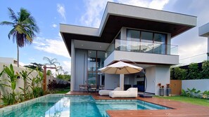 Una casa moderna de dos pisos con piscina, mobiliario exterior y abundante vegetación.