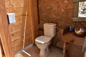 A small, rustic bathroom with a wooden door, a white toilet, a stone wall, and a wooden shelf with a metal bowl.