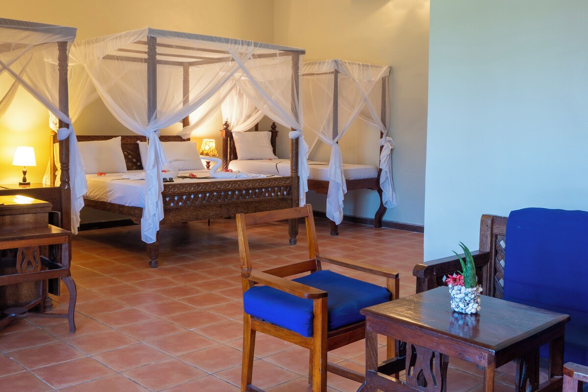 A four-poster bed with a canopy, a wooden chair with a blue cushion, and a small table with a decorative plant.