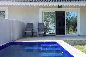 Zona de piscina con dos sillas de madera y una mesa pequeña, un suelo de baldosas y un edificio con fachada blanca y detalles decorativos.