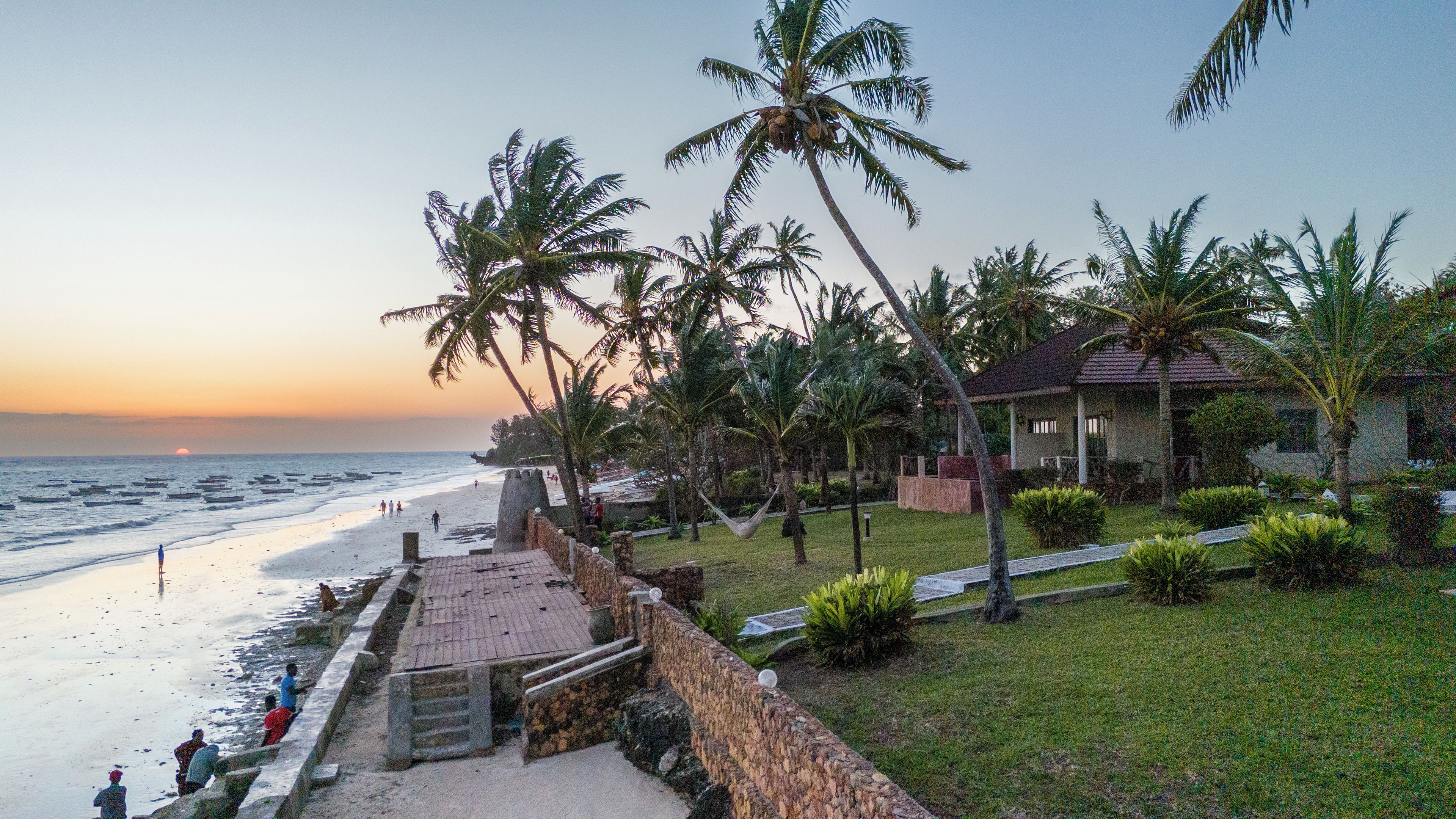 A beachfront property with a stone pathway leading to the beach, palm trees, and a small building with a tiled roof.