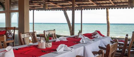 A dining area with tables set for two, overlooking the sea, featuring wooden chairs and a red tablecloth.