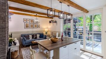 A modern kitchen with a wooden ceiling, pendant lights, a dining area with a blue sofa, and a view of the outdoors through large windows.
