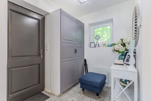 En gang med en grå dør, en grå garderobe, en blå bænk, et hvidt konsolbord med hat og lampe, samt et vindue med udsigt til grøn vegetation.
