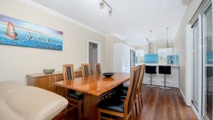 A modern dining area with a wooden table and chairs, a painting of sailboats on the wall, and a kitchen in the background.