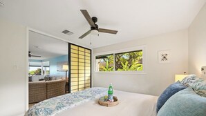 A bedroom with a bed, a ceiling fan, a window with a view of greenery, and a wicker basket with a bottle on the bed.
