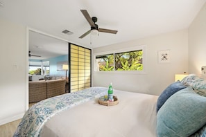 Une chambre avec un lit, un ventilateur de plafond, une fenêtre donnant sur un paysage verdoyant et un panier en osier contenant une bouteille, posé sur le lit.