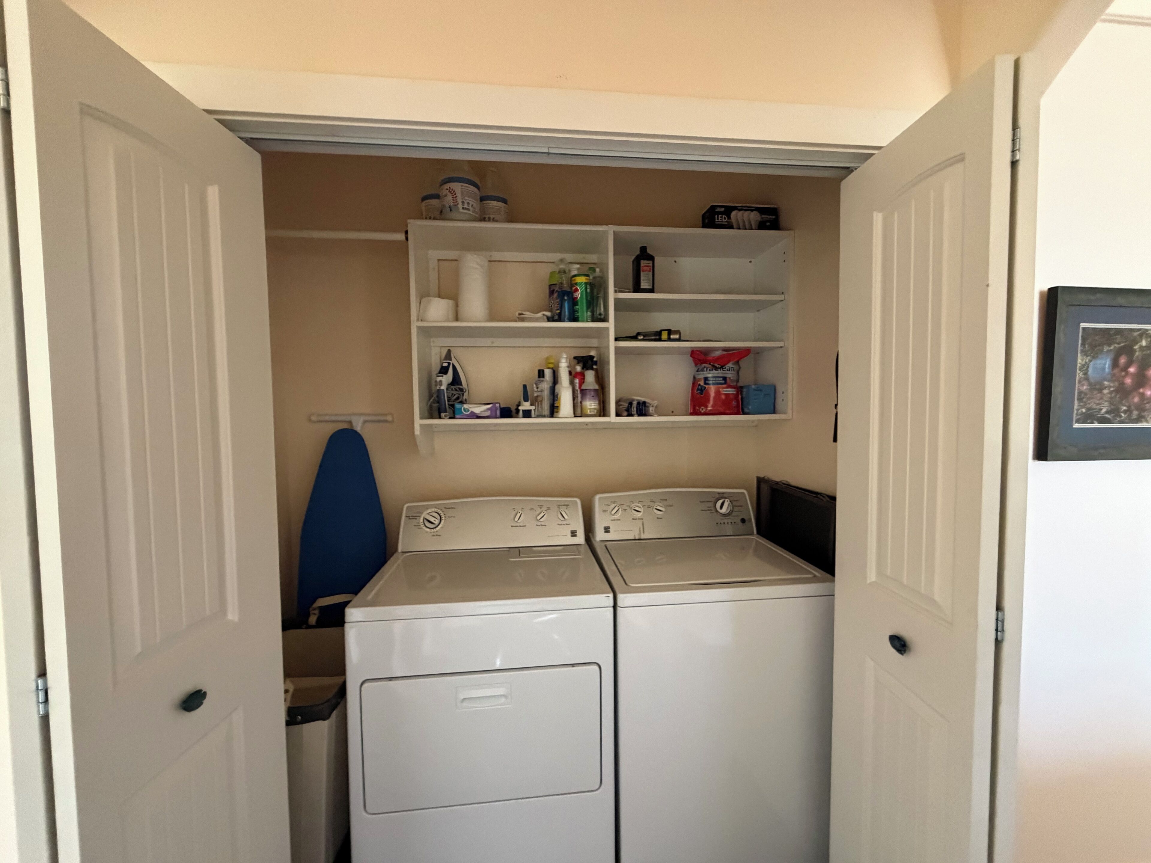 A laundry room with two washing machines, a shelf with various items, and a blue ironing board.