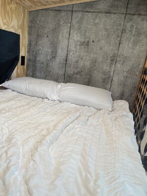 A bed with a white quilt and pillows against a concrete headboard in a room with wooden walls. - Unplug and unwind in this charming loft cabin nestled on the water’s edge. (Odessa)