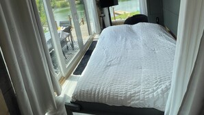 Une chambre à coucher avec un lit, un ventilateur de plafond, une télévision et une vue sur un lac, visible à travers de grandes fenêtres.