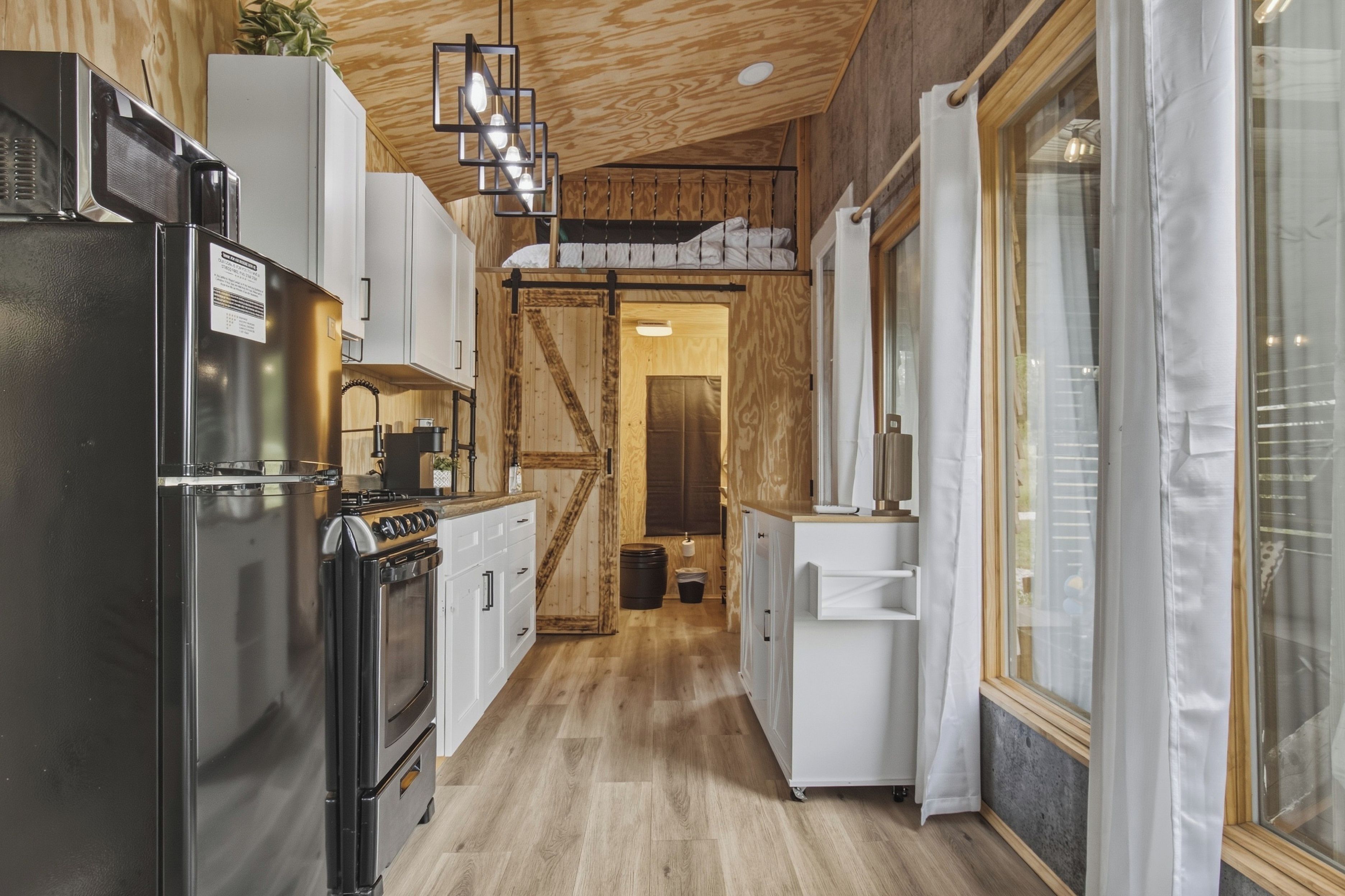 A narrow kitchen with wooden flooring, white cabinetry, a black refrigerator, and a hanging light fixture.