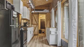 A narrow kitchen with wooden flooring, white cabinetry, a black refrigerator, and a hanging light fixture.