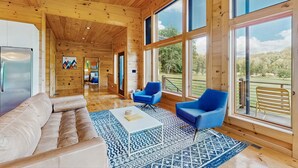 A spacious living room with a wooden ceiling, large windows, a beige sofa, blue armchairs, and a patterned rug.