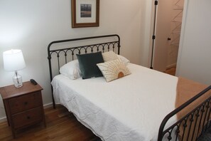 Een netjes opgemaakt bed met een witte dekbedovertrek en meerdere kussens, een nachtkastje met een lamp en afstandsbediening, en een lijst met een foto aan de muur.