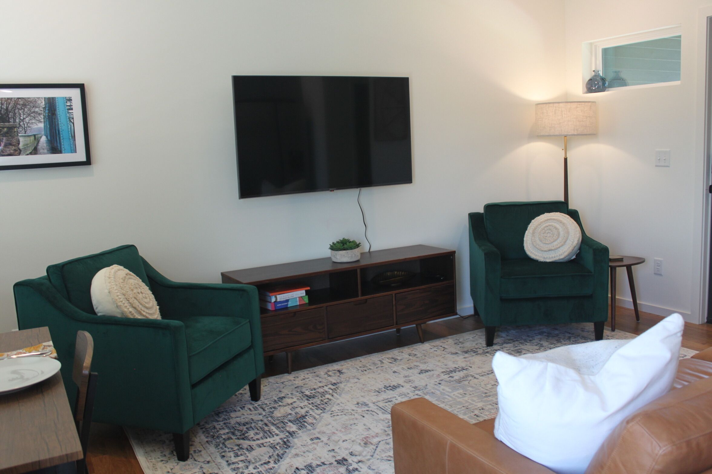 A living room with a flat-screen TV mounted on the wall, a green armchair, a brown leather sofa, and a small side table with a lamp.