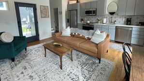 A modern living room with a sofa, coffee table, and a green armchair. A kitchen with grey cabinets and stainless steel appliances is visible in the background. - Live in Southside By Trails & Downtown + Parking (Chattanooga)