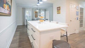 A kitchen with a central island, a dining area with chairs, and a hallway leading to a door. - Fire pit - 6 Min to North Hills - Screened Porch (Raleigh)