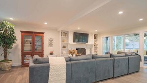 A modern living room with a grey sectional sofa, a wooden cabinet, a TV, and a large window. - Fire pit - 6 Min to North Hills - Screened Porch (Raleigh)
