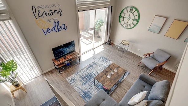 A modern living room with a sofa, coffee table, and TV. There's a large window with blinds, a potted plant, and a wall clock. - Vintage Stay, 1BR Old Town Scottsdale Retreat (Scottsdale)
