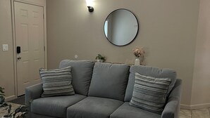 Un salon avec un canapé gris, deux coussins à motifs, un miroir rond et une petite table sur laquelle sont posés des vases.