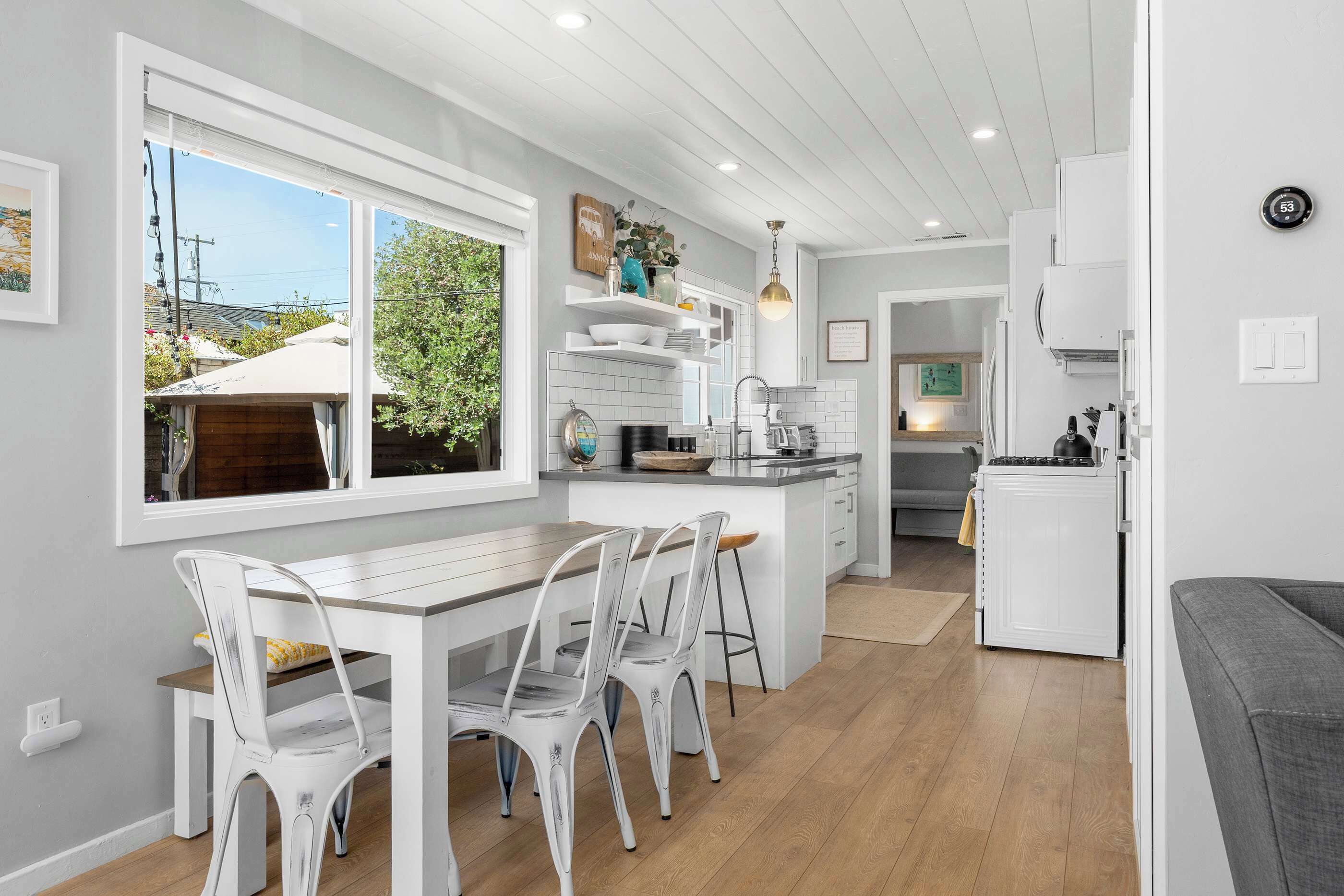 Une cuisine moderne avec des meubles blancs, un coin repas avec des chaises blanches et une vue sur l’extérieur grâce à de grandes fenêtres.