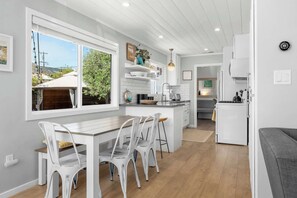 A modern kitchen with white cabinetry, a dining area with white chairs, and a view of the outdoors through large windows.