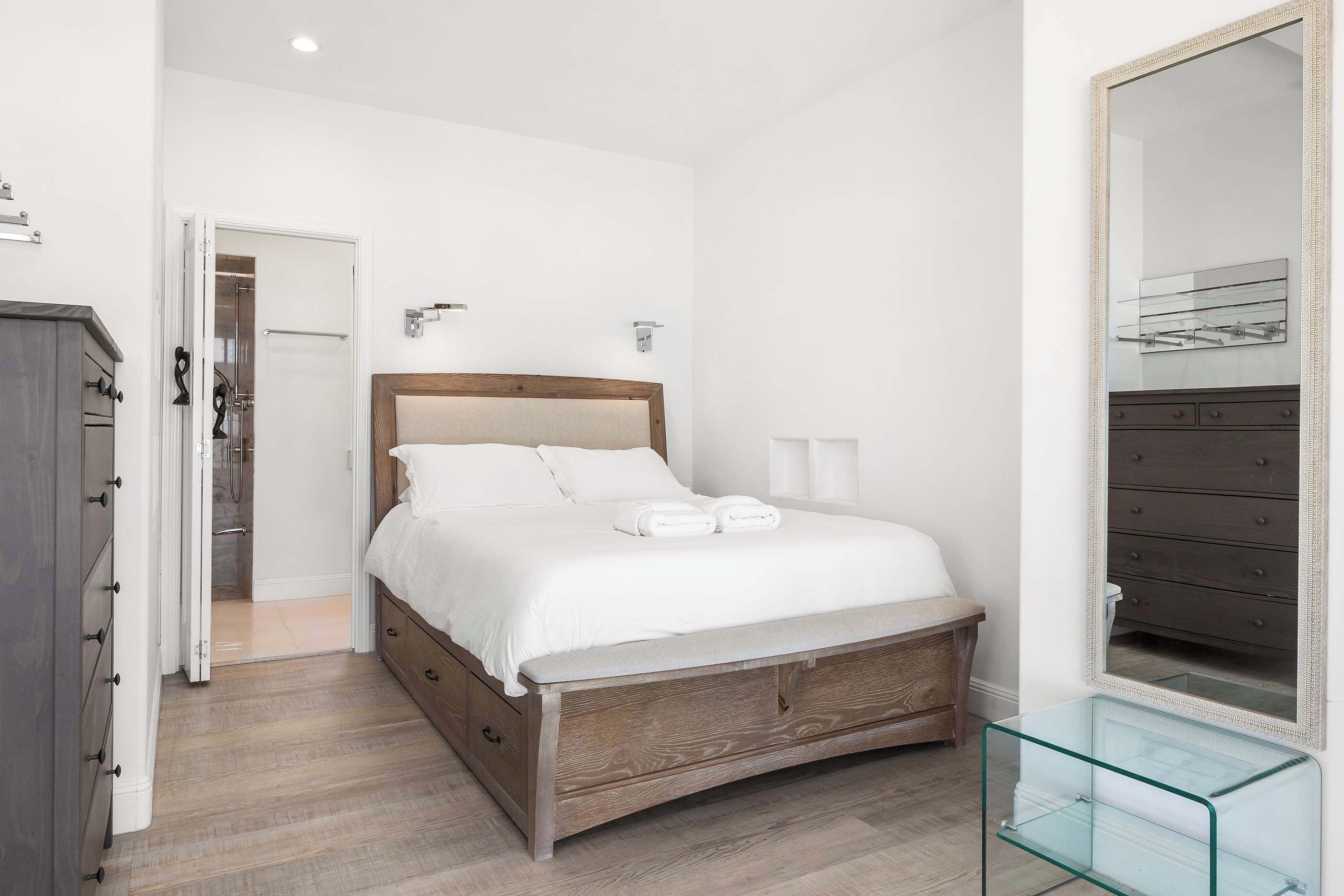 A bedroom with a wooden bed, a dresser, a glass side table, and a mirror on the wall.