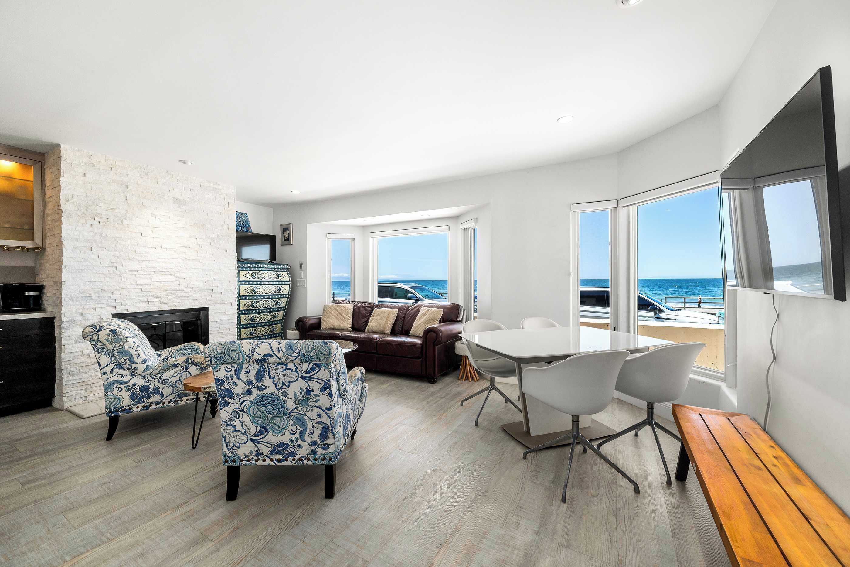 Un vaste espace de vie agrémenté d’un mur en briques blanches, d’une cheminée, d’un canapé, d’une table à manger avec des chaises et de grandes fenêtres offrant une vue sur la mer.