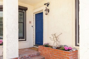 A blue door with a decorative border, a small brick planter with flowers, and a wall-mounted light fixture. - 3 Bedroom Rio Del Mar Home - Steps to Beach! (Aptos)