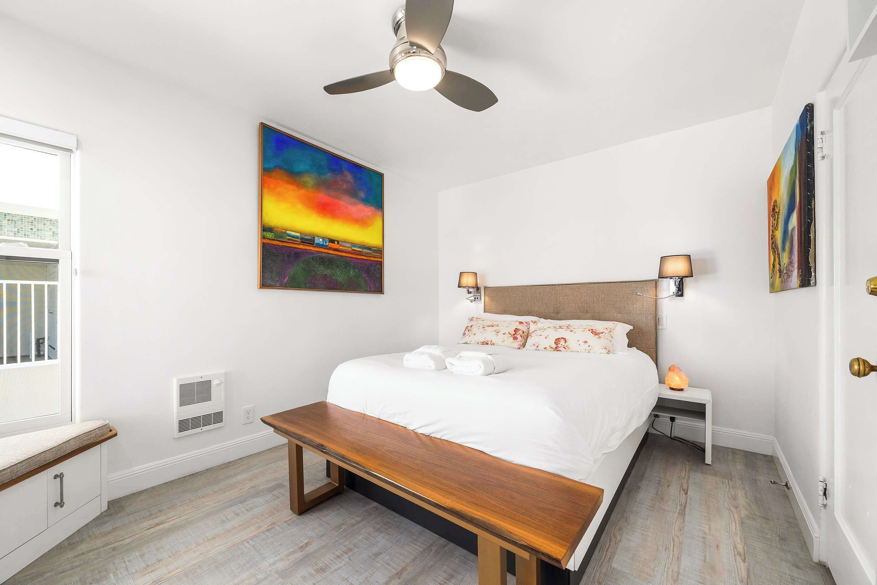 A bedroom with a bed, bedside lamps, a ceiling fan, a bench, and artwork on the walls.