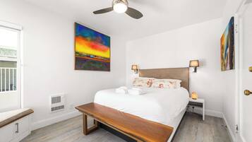 A bedroom with a bed, bedside lamps, a ceiling fan, a bench, and artwork on the walls.