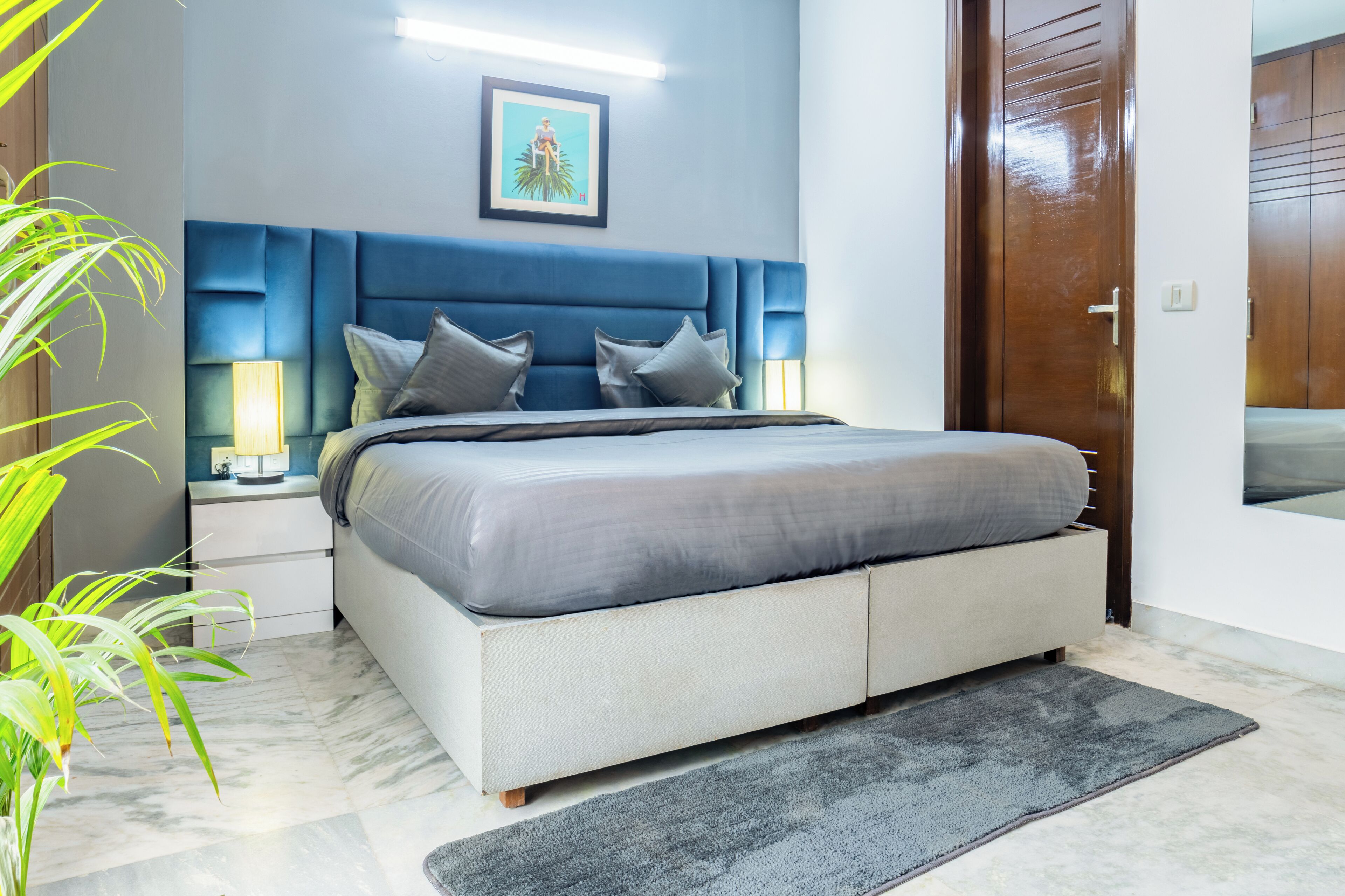 A modern bedroom with a large bed, two bedside tables with lamps, a blue headboard, a framed picture on the wall, and a wooden door.