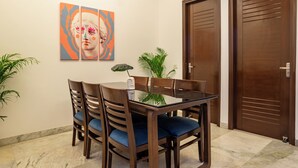 A dining area with a glass table, wooden chairs with blue cushions, and a painting on the wall.