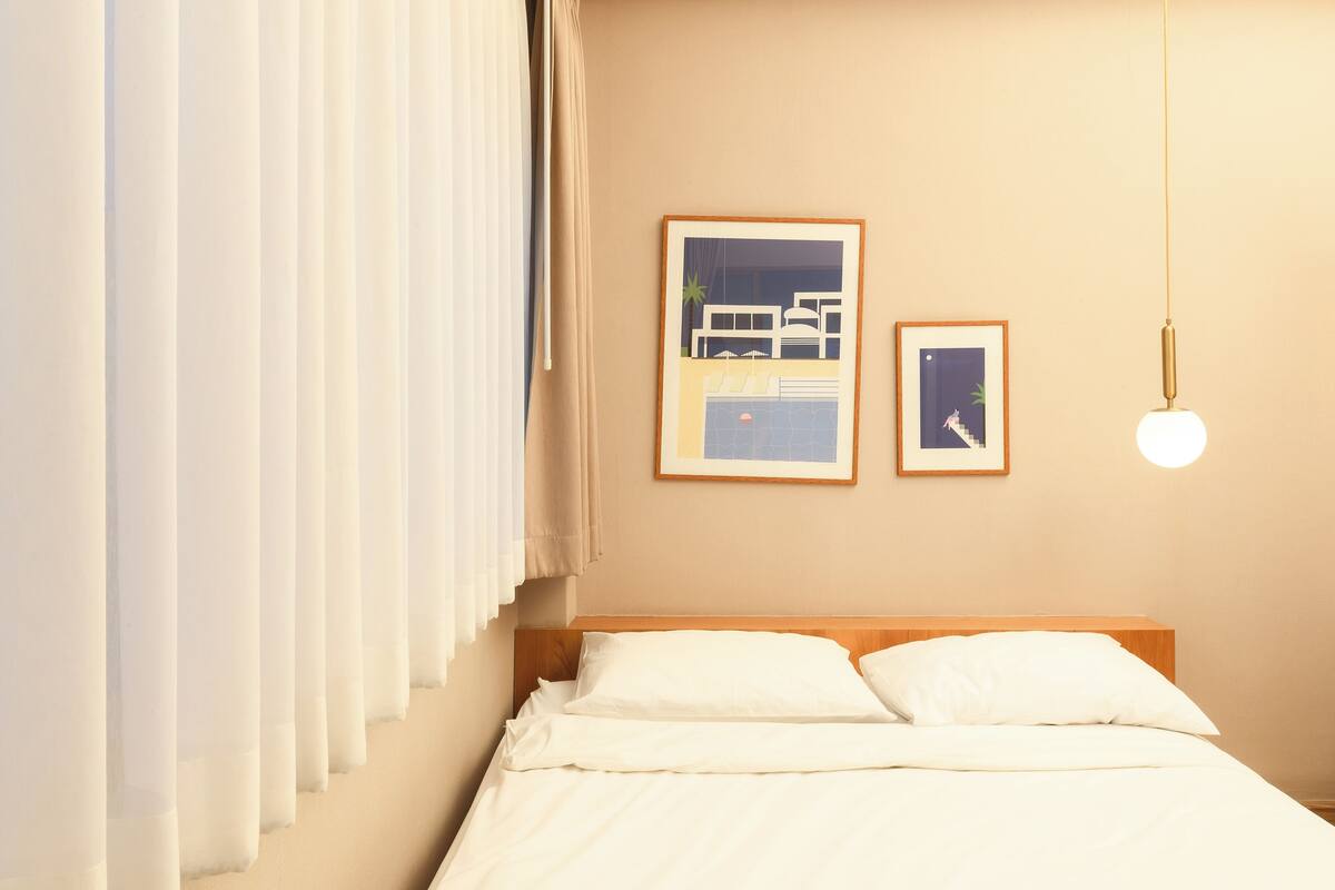 Un lit bien fait, avec du linge de lit blanc, dans une pièce aux murs de couleur beige. Deux tableaux encadrés sont accrochés au mur, l’un représentant une scène de plage et l’autre un escalier.