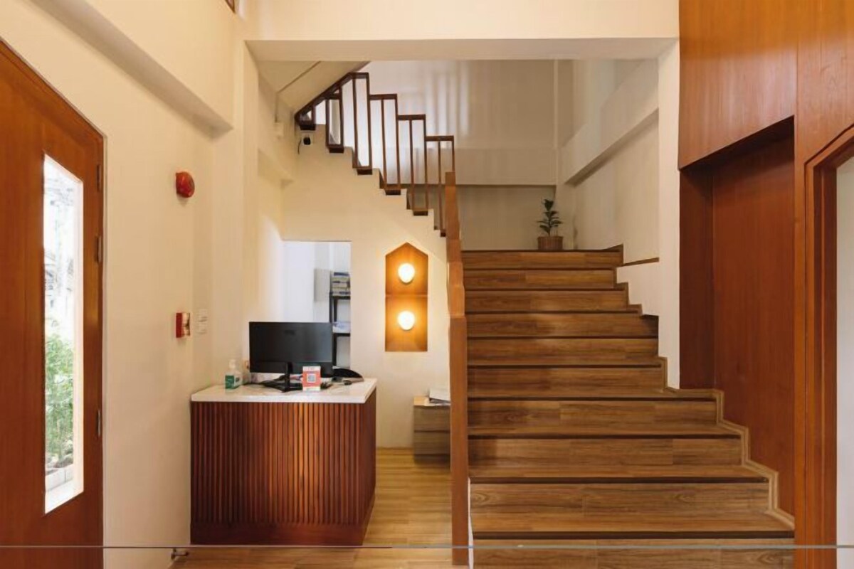 A staircase with wooden steps leading to an upper floor, a reception desk with a computer and a small plant, and a wall-mounted light fixture.