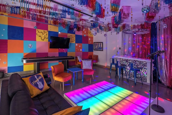 A vibrant room with a colorful checkered wall, a flat-screen TV, a sofa with a pillow, a bar area, and a disco ball.