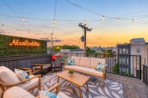 An outdoor seating area with a sofa, chairs, and a table, overlooking a cityscape at sunset.