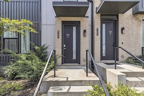 A modern building with two black doors marked with numbers 516-5 and 516-6, a concrete staircase with metal railings, and a small garden area with shrubs.
