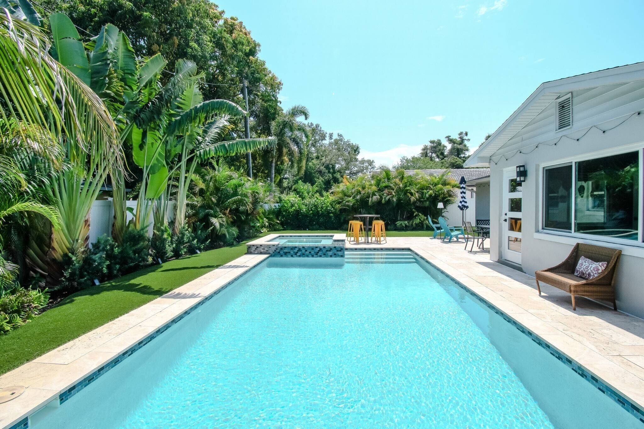 A rectangular swimming pool with a tiled edge, surrounded by lush greenery and a white building with large windows.